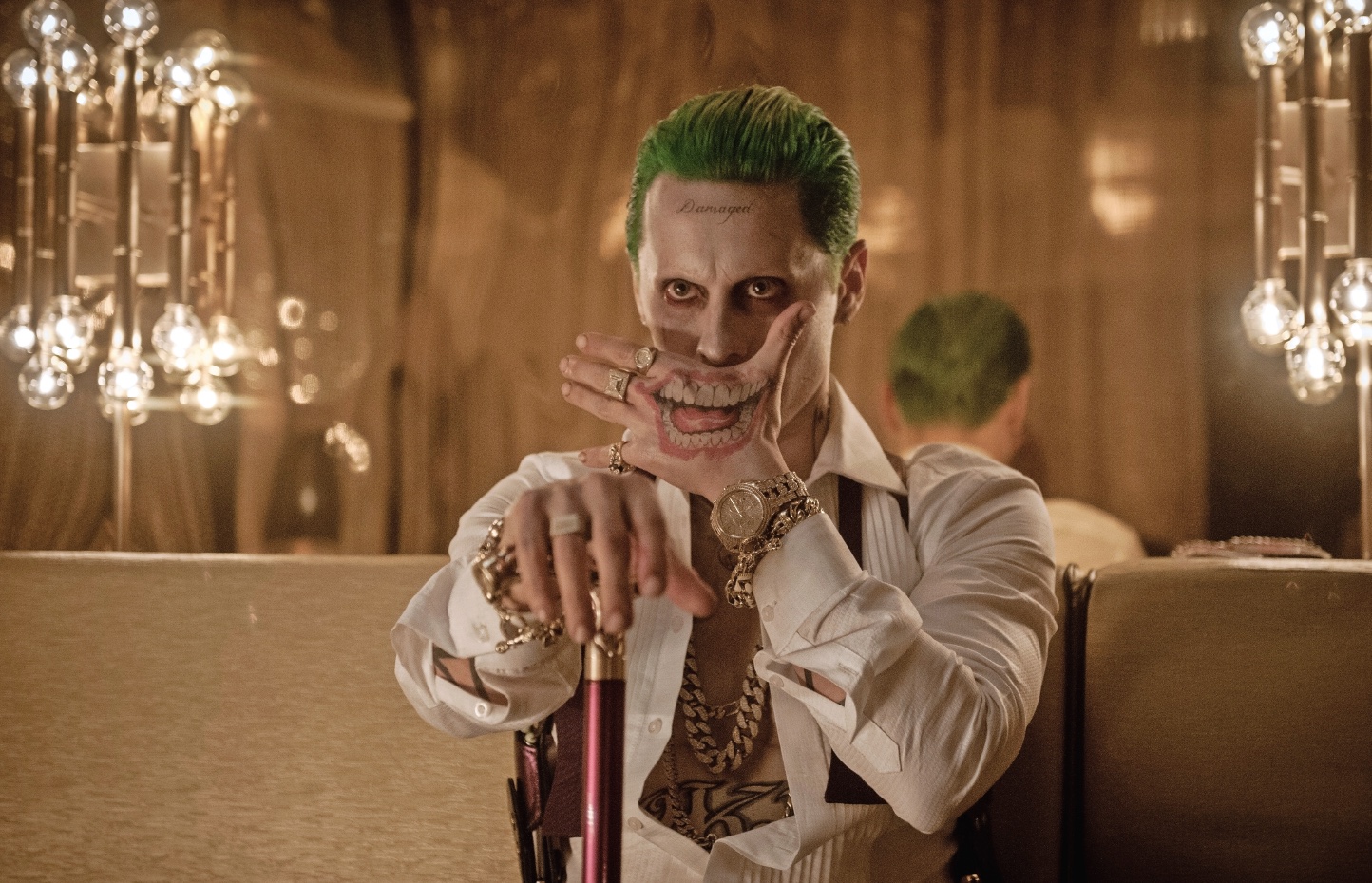 Close-up portrait of actor Jared Leto in his portrayal of the Joker from the movie "Suicide Squad." He has green hair, damaged tattoos, and a purple coat, with a menacing grin on his face.